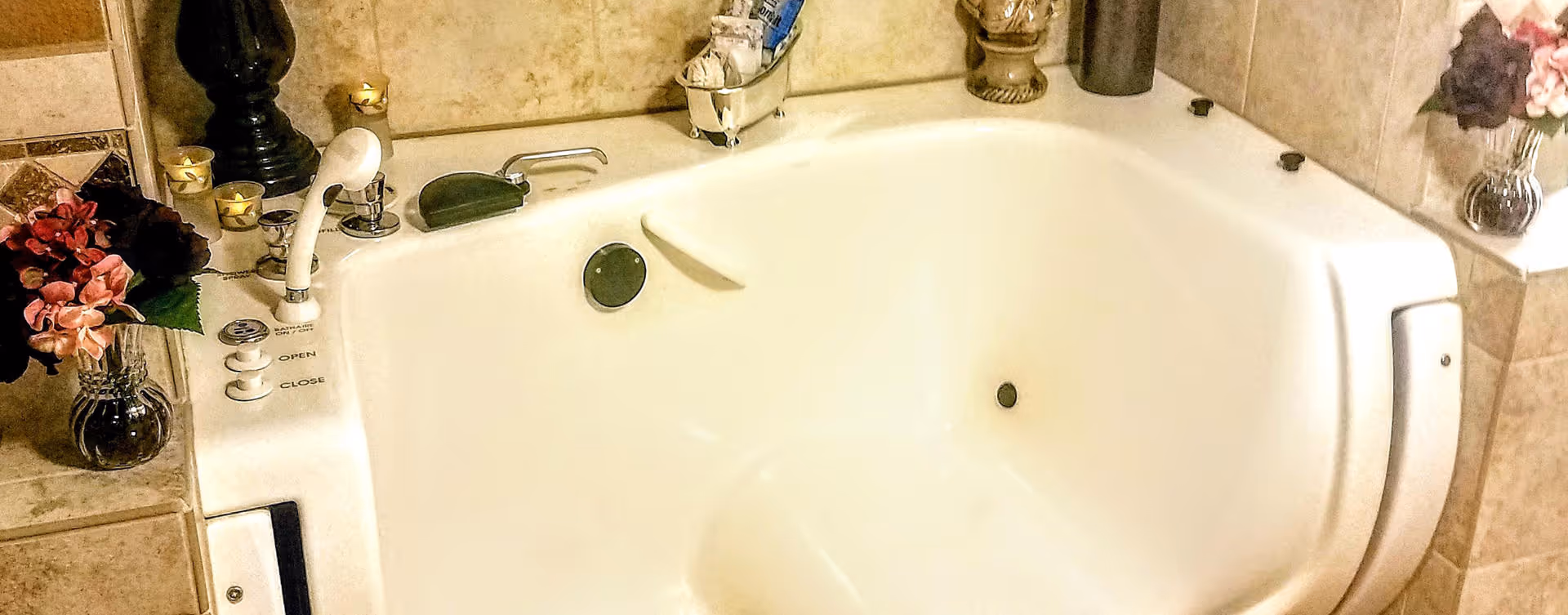 A white corner bathtub with built-in controls and a handheld showerhead, surrounded by beige tiled walls and decorative items including candles and flower arrangements in vases.