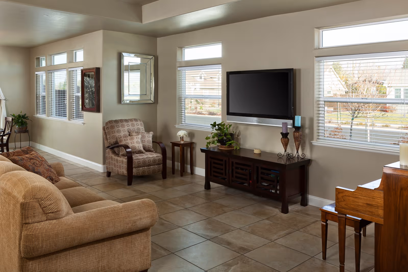 A cozy living room with a beige couch, a patterned armchair, a wooden TV stand with a flat-screen TV mounted above it, and a piano with a bench. The room has large windows with white blinds letting in natural light, beige walls, and tiled flooring. There are decorative items including plants, candles, and a wall mirror.