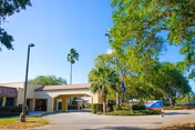 Exterior view of Heritage Park Health Center showing the entrance driveway with a covered drop-off area, surrounded by green trees and landscaping under a clear blue sky.