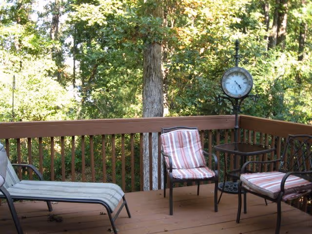 Wooden outdoor deck with cushioned chairs, a lounge chair, a small table and a standing clock surrounded by trees.