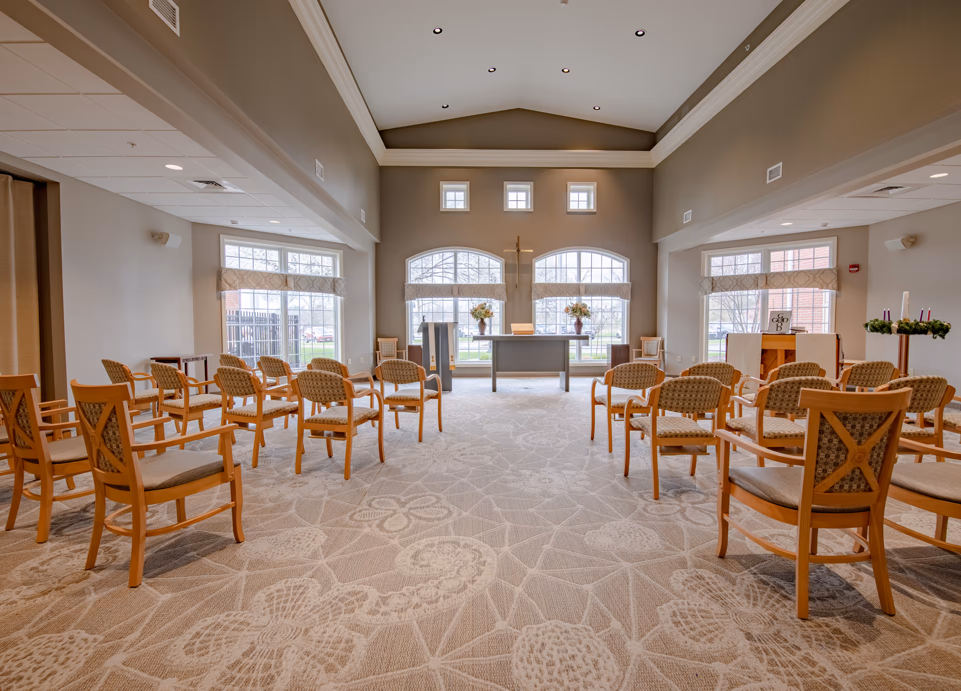 A bright and spacious chapel or worship room with rows of wooden chairs arranged facing a small altar. The altar is positioned in front of three large windows with floral arrangements on either side and a cross mounted on the wall above. The room has a high ceiling with recessed lighting and a patterned carpet with floral designs.