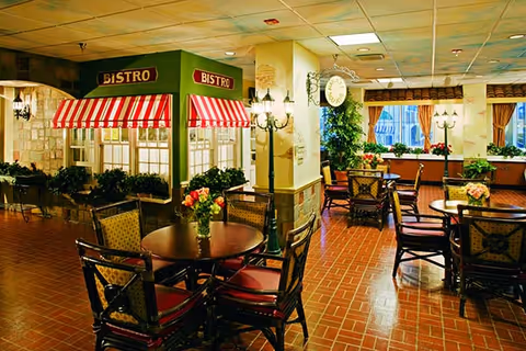 Interior view of a retirement community dining area featuring multiple round tables with chairs. The space includes a decorative bistro setup with red and white striped awnings and green signage labeled 'BISTRO'. The room has warm lighting, brick flooring, potted plants, and large windows with curtains allowing natural light to enter.