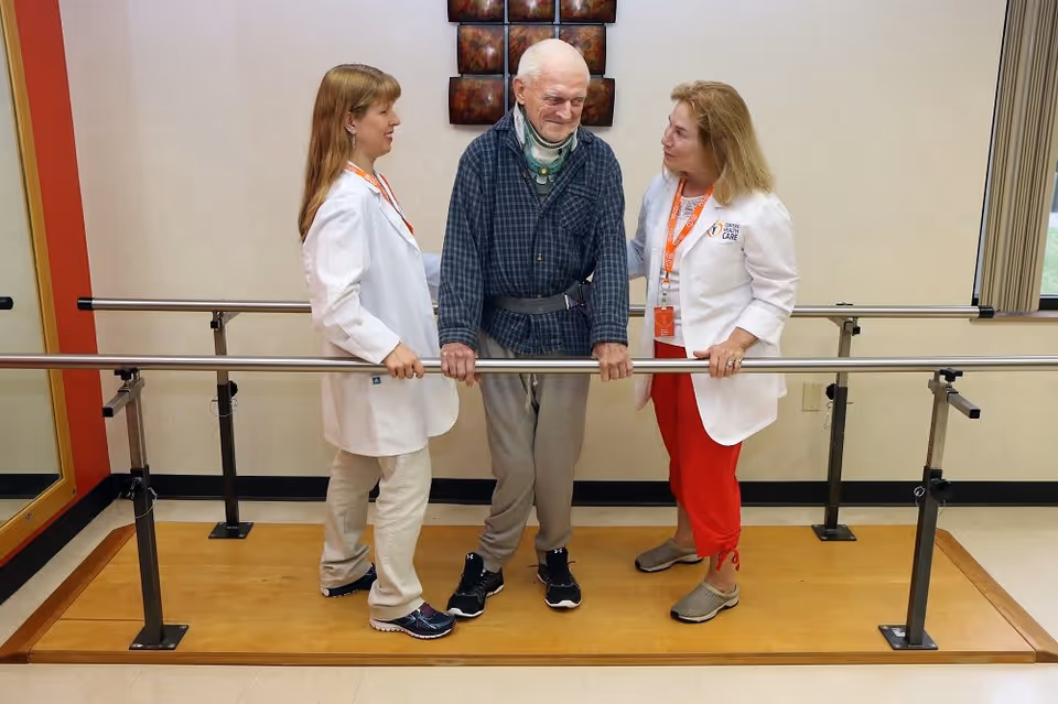 An elderly man wearing a neck brace and support belt is standing between two female healthcare professionals in white coats. They are in a rehabilitation room with parallel bars used for physical therapy. The man is holding onto the bars while the women support and encourage him.