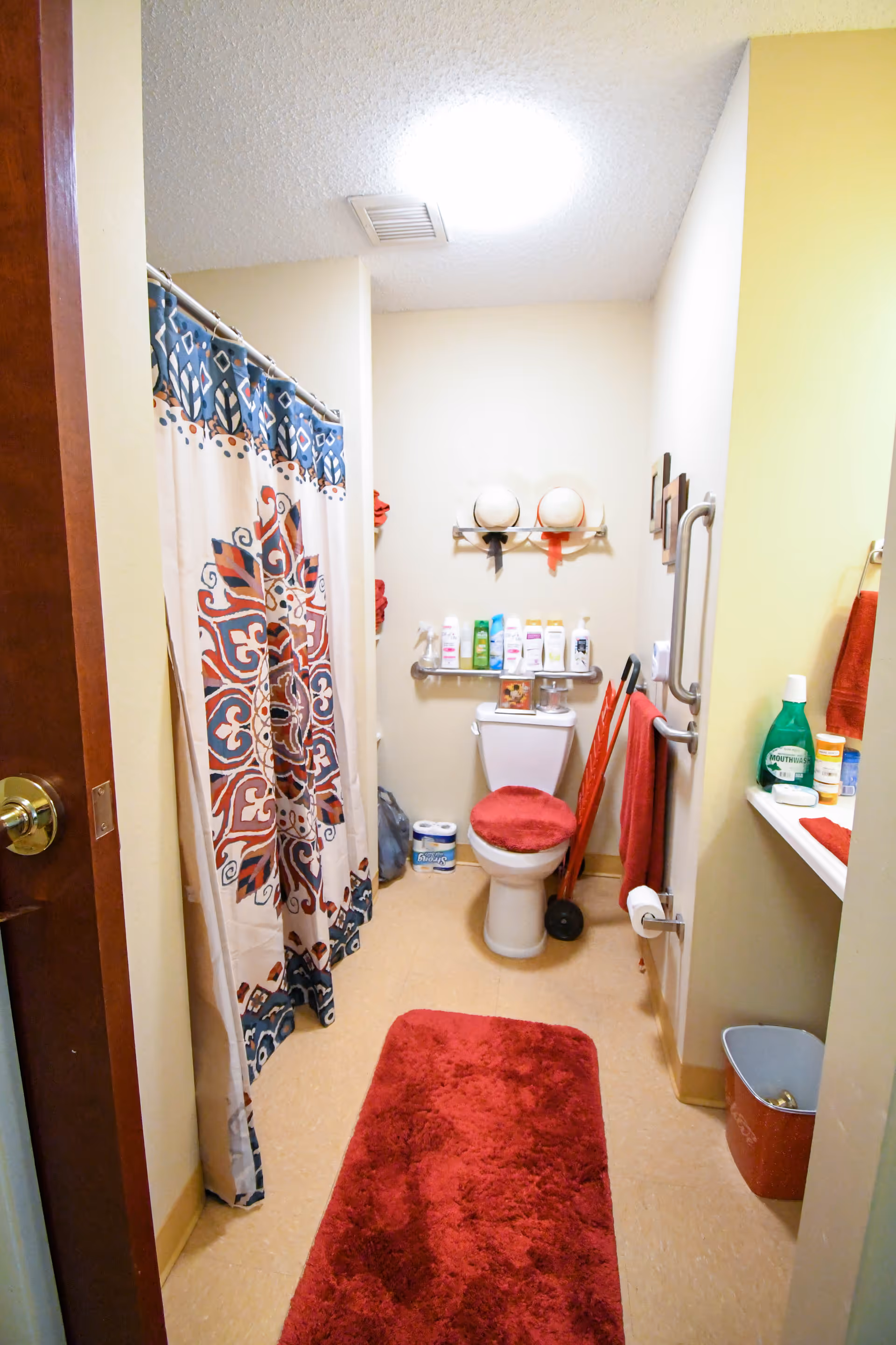 A small bathroom with a toilet, a shower with a patterned curtain, and a red rug on the floor. The toilet has a red cover on the seat and a shelf above it holding various toiletries. There are red towels hanging on the wall and a grab bar next to the toilet. A small trash bin and cleaning supplies are visible on the right side.