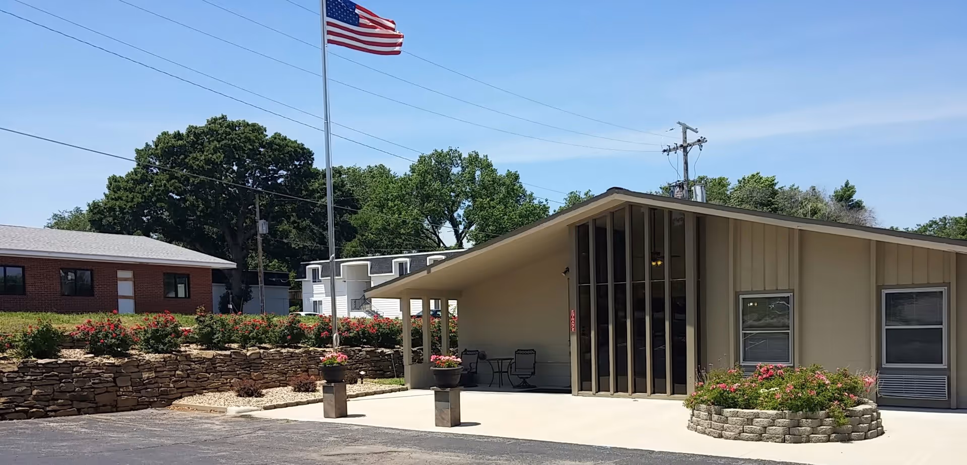 Exterior view of a senior living facility building with a slanted roof, large vertical windows, and a small patio area with chairs and a table. There are flower beds with pink flowers and a tall flagpole with an American flag waving. Other buildings and trees are visible in the background under a clear blue sky.
