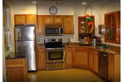 A kitchen area with wooden cabinets, stainless steel refrigerator, oven, microwave, and dishwasher. There is a clock on the wall above the cabinets and a small decorative shelf with a plant and other items.