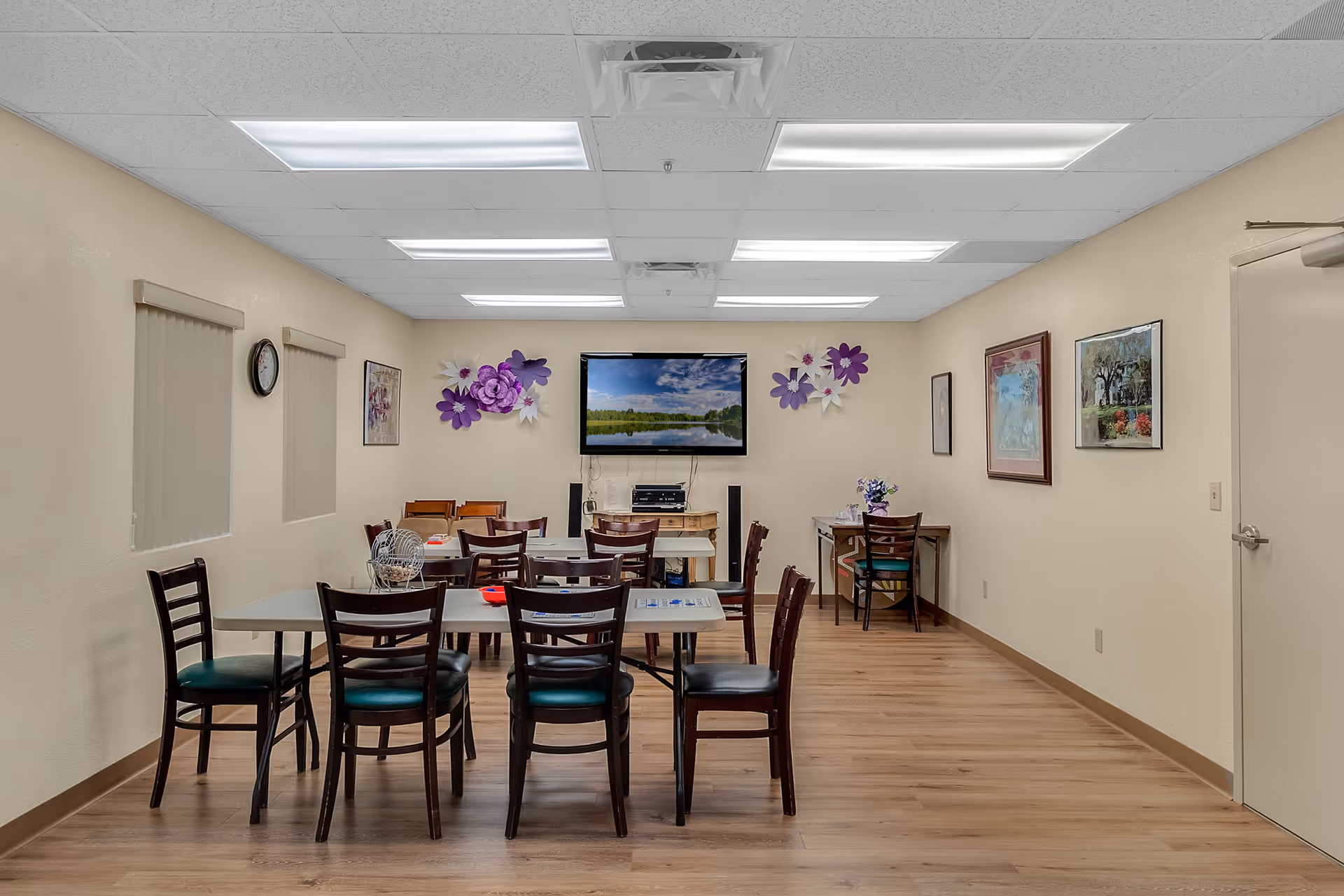 A well-lit room with several tables and chairs arranged for group seating. The walls are decorated with framed pictures and purple floral wall art. A flat-screen TV is mounted on the far wall above a small table with electronic equipment. The floor is wooden, and the ceiling has fluorescent lighting panels.
