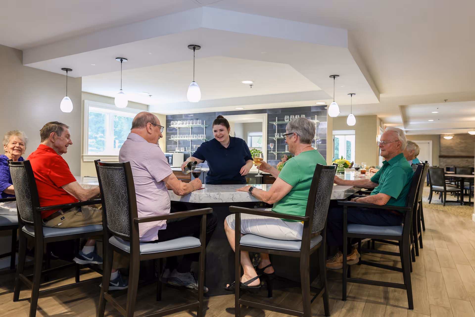 A group of elderly people sitting around a large marble table in a well-lit dining area, smiling and enjoying drinks served by a staff member. The room has modern pendant lights and large windows letting in natural light.