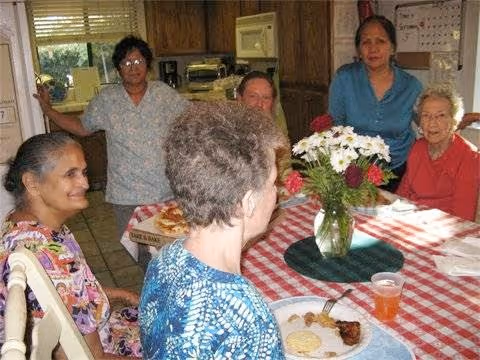 A group of six elderly women gathered around a dining table with a red and white checkered tablecloth. There is a vase with white and red flowers in the center of the table. Some women are seated and some are standing in a kitchen setting with wooden cabinets and a microwave in the background. Plates with food and a drink are visible on the table.