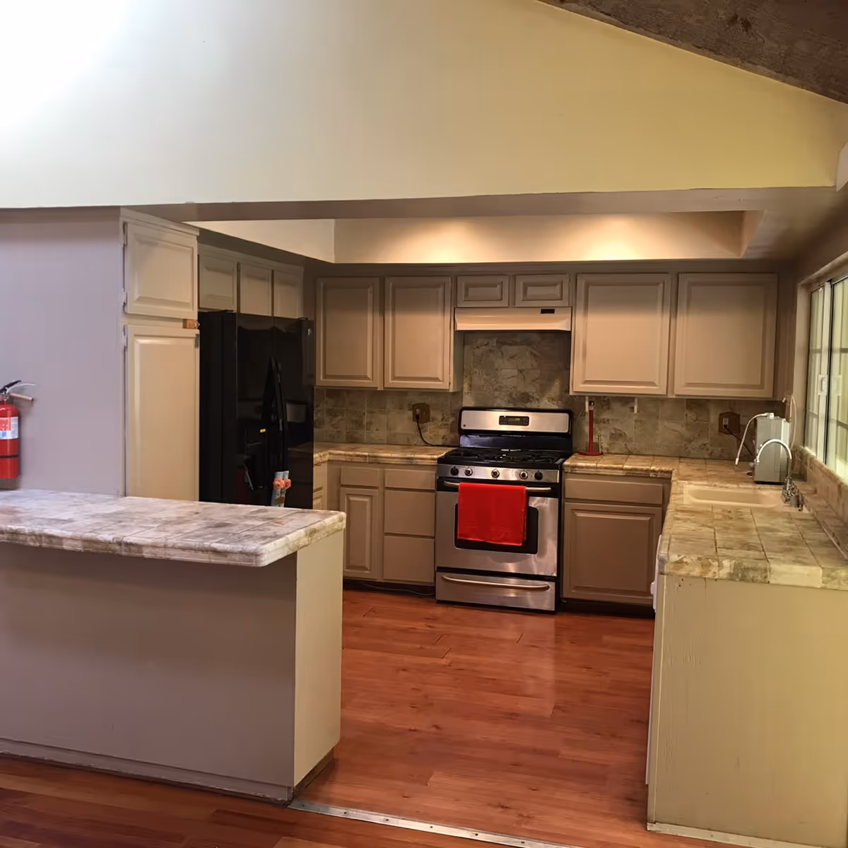 Interior view of a kitchen with beige cabinets, a stainless steel stove with a red towel hanging on the handle, a black refrigerator, tiled countertops, and wooden flooring. There is a fire extinguisher mounted on the wall to the left and a window on the right side above the sink.