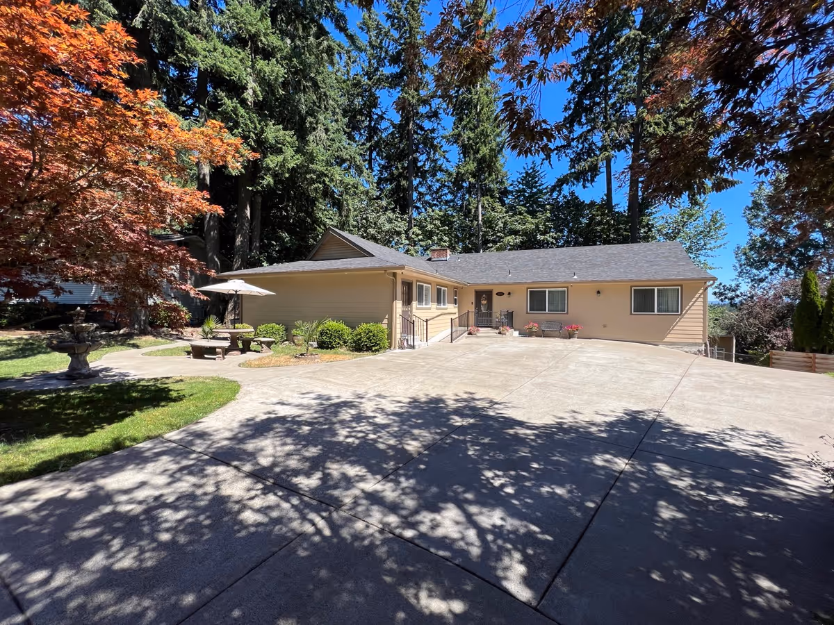 A single-story beige house with a large concrete driveway in front, surrounded by tall trees and greenery. There is a small patio area with a round table, benches, and an umbrella on the left side. The sky is clear and blue, and shadows of tree branches fall on the driveway.