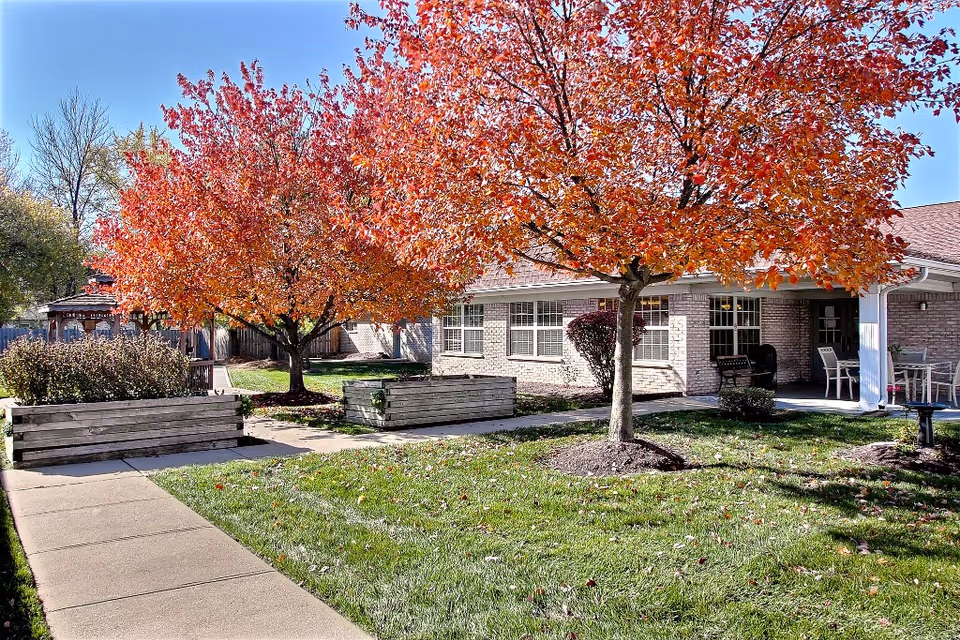Courtyard with autumn trees, raised planters, a sidewalk, and the brick front of a senior living building with patio seating.