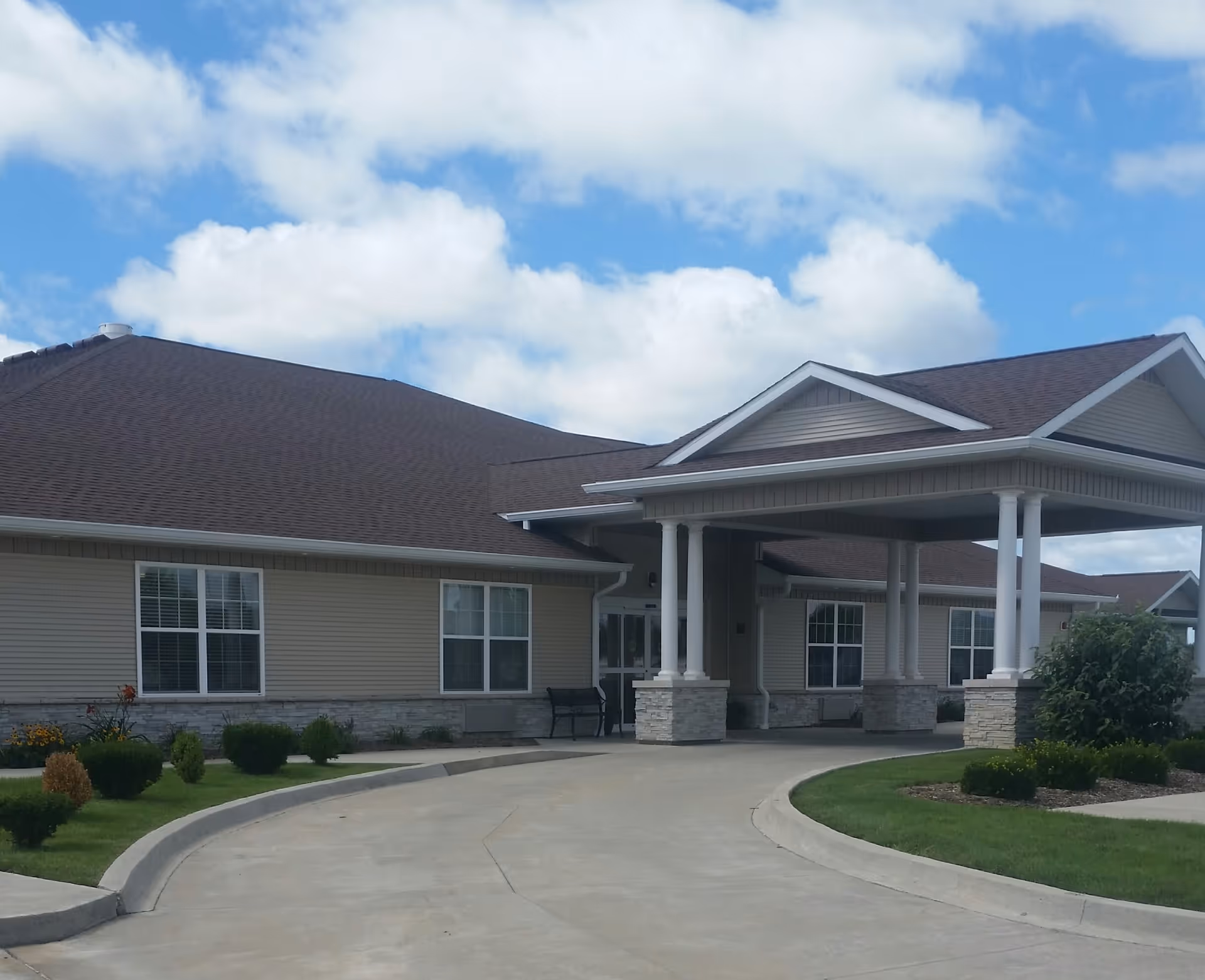 Exterior view of a single-story building with beige siding and a dark brown roof under a partly cloudy sky. The building features a covered entrance supported by white columns and a curved driveway leading up to it. There are small bushes and landscaped greenery around the building.
