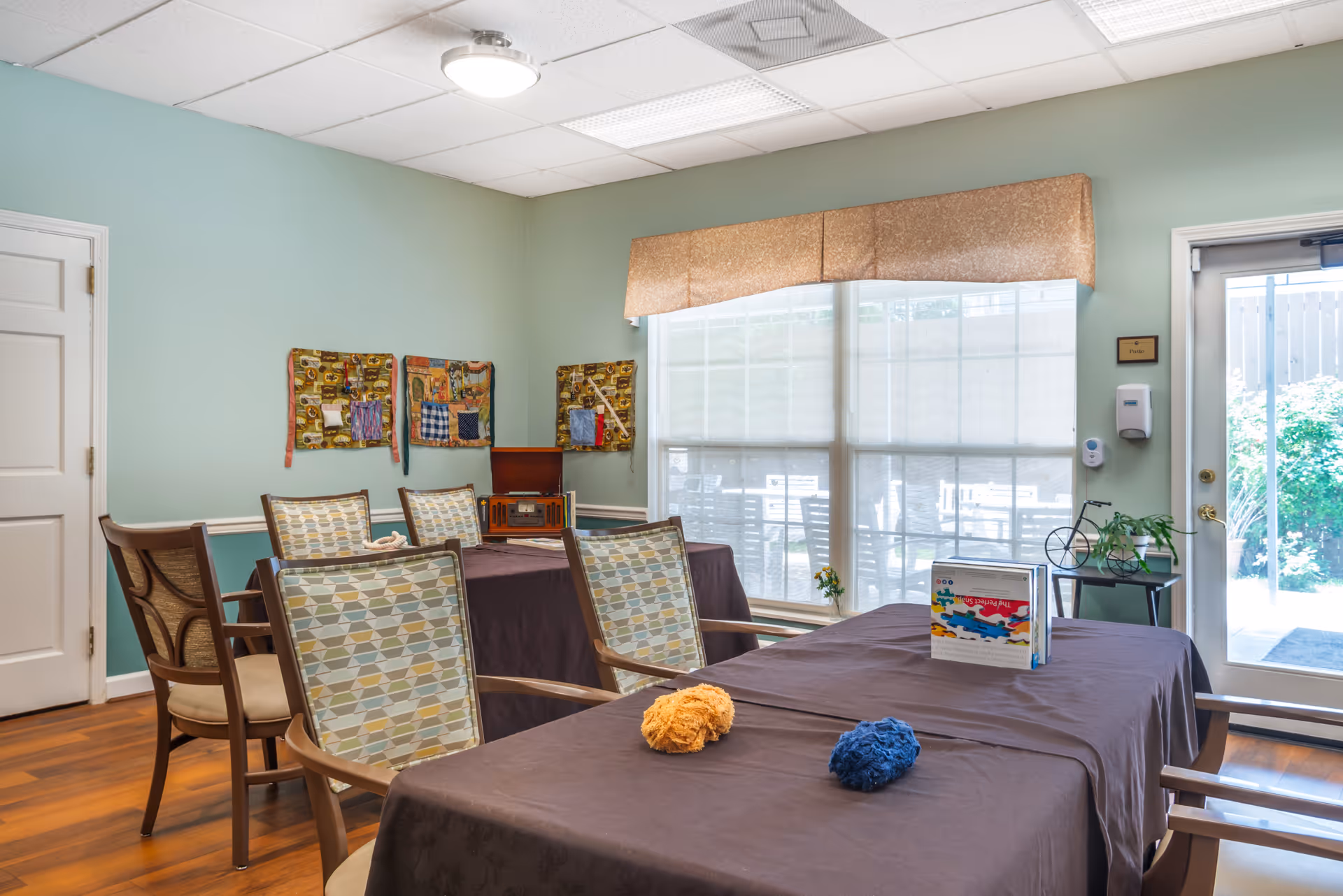 Well-lit communal activity room with tables covered in dark tablecloths, patterned chairs, craft supplies and large windows leading to an outdoor area.