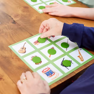 Hands place green tokens on a grid of illustrated cards on a wooden table.