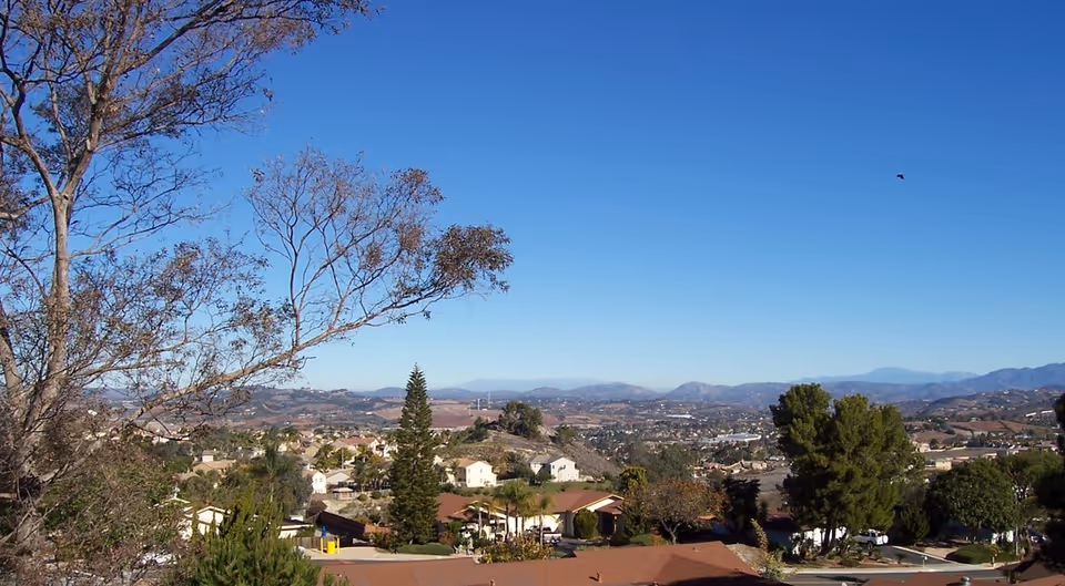 A scenic view of a suburban neighborhood with houses, trees, and hills in the background under a clear blue sky.