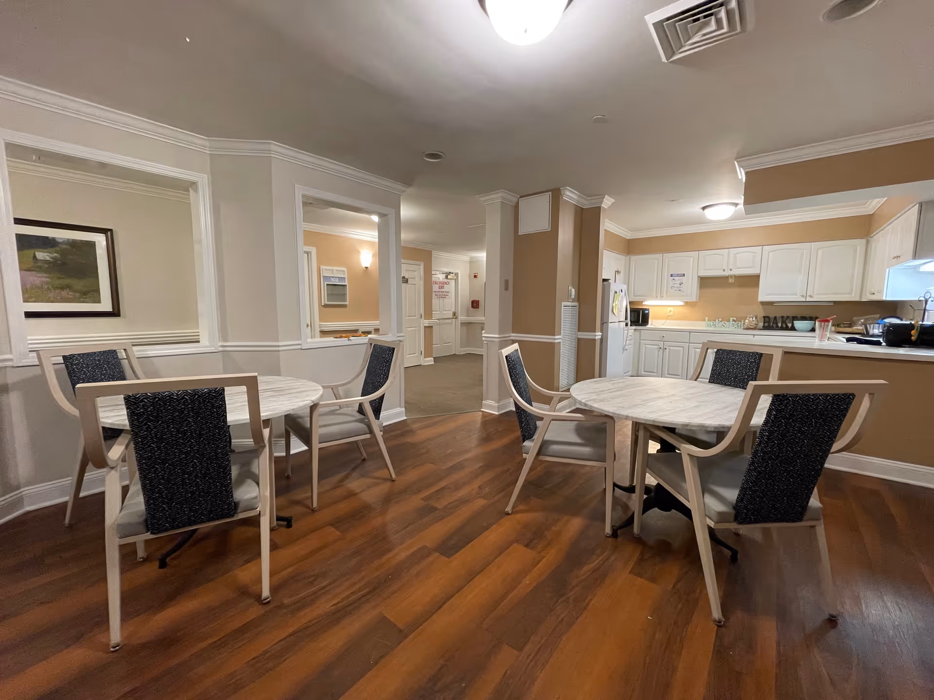 Interior view of a senior living facility dining area with two round tables, each surrounded by four chairs with dark patterned backs and light seats. The room has wood flooring and beige walls with white trim. In the background, there is a kitchen area with white cabinets, a refrigerator, and a countertop with various items on it. The space is well-lit with ceiling lights.