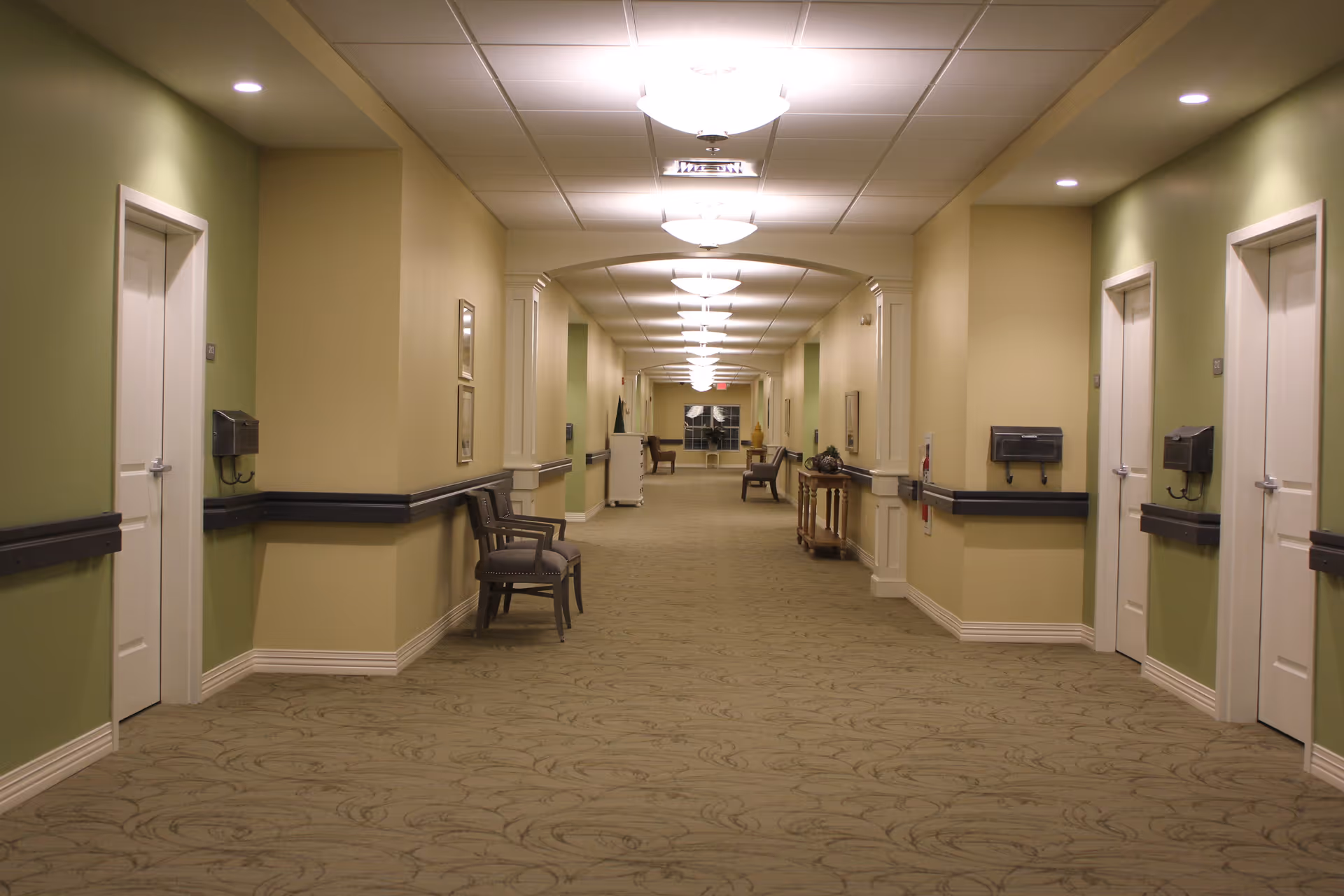 A long, well-lit hallway in a senior living facility with beige and green walls, carpeted floor, multiple closed white doors on both sides, chairs placed along the walls, and ceiling lights evenly spaced down the corridor.