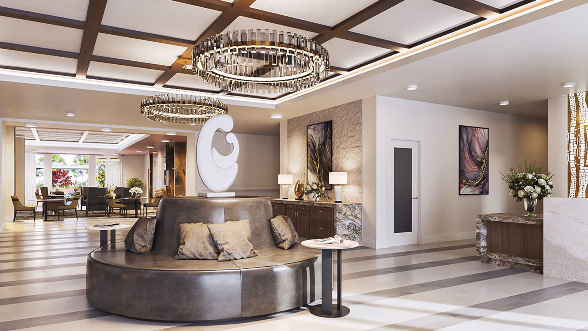 A modern and elegant assisted living facility common area with a circular leather seating arrangement in the center, decorative pillows, two round side tables, and a large abstract sculpture. The room features stylish chandeliers, contemporary artwork on the walls, a sideboard with lamps and floral arrangements, and a dining area with tables and chairs visible in the background near large windows.