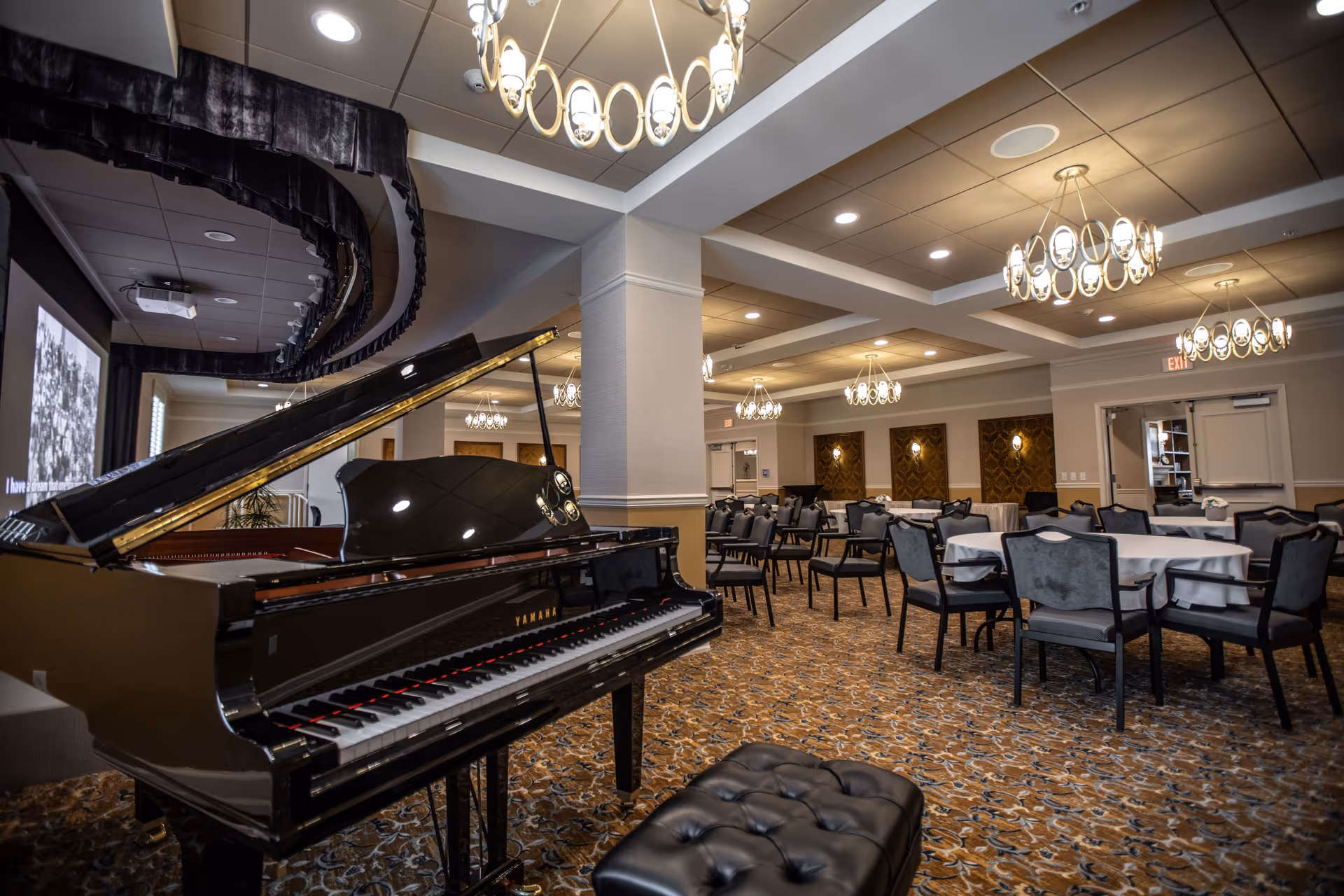 Spacious dining/event room with a grand piano, round tables and chairs under multiple chandeliers.