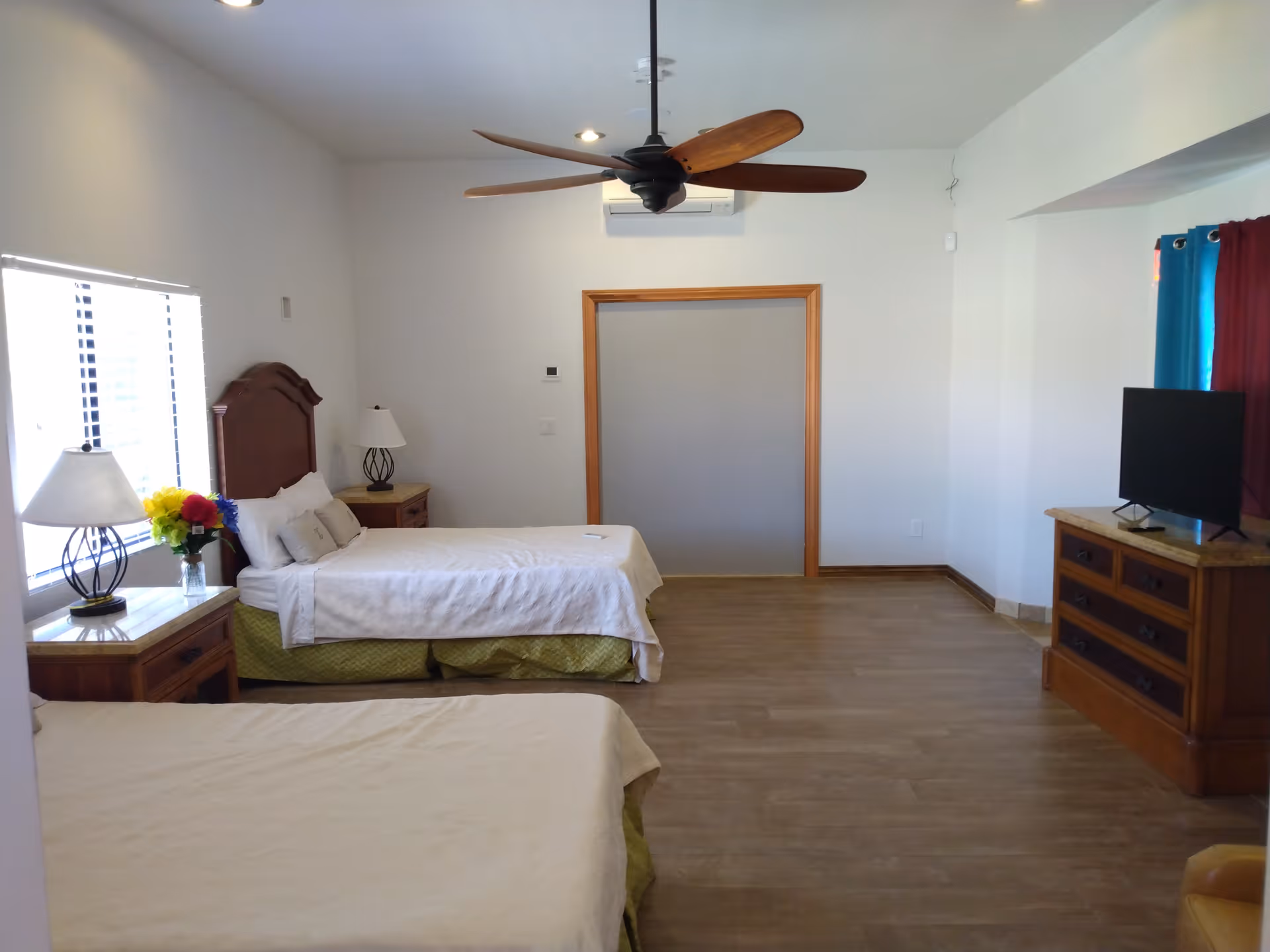 A spacious bedroom with two beds, each with white bedding and green bed skirts. There are two bedside tables with lamps and a vase of colorful flowers on one table. A ceiling fan with wooden blades is mounted on the ceiling. A flat-screen TV sits on a wooden dresser near a window with blue and red curtains. The room has wooden flooring and white walls.