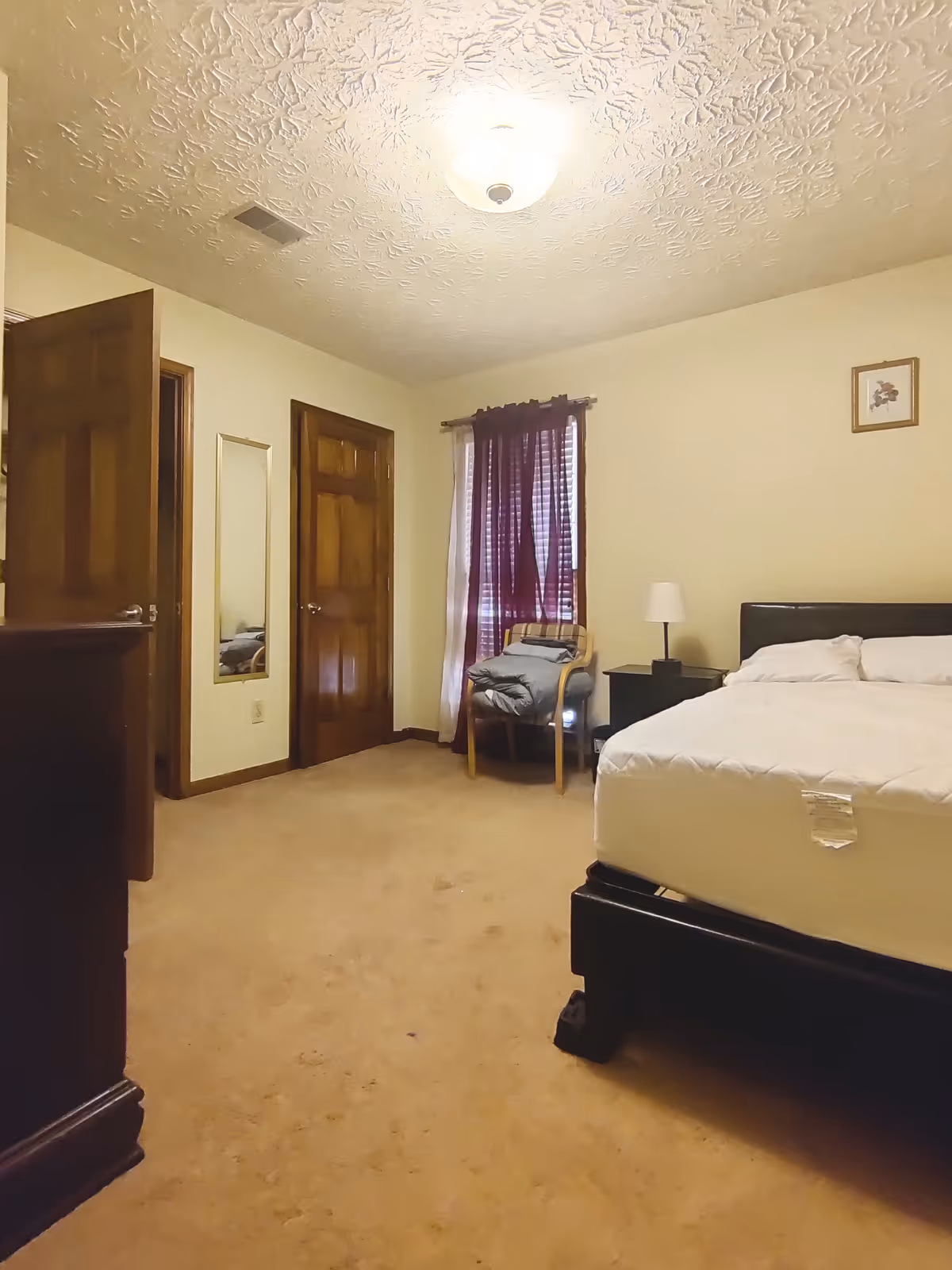 Furnished bedroom with a bed, nightstand and lamp, a chair by a curtained window, and two wooden doors.