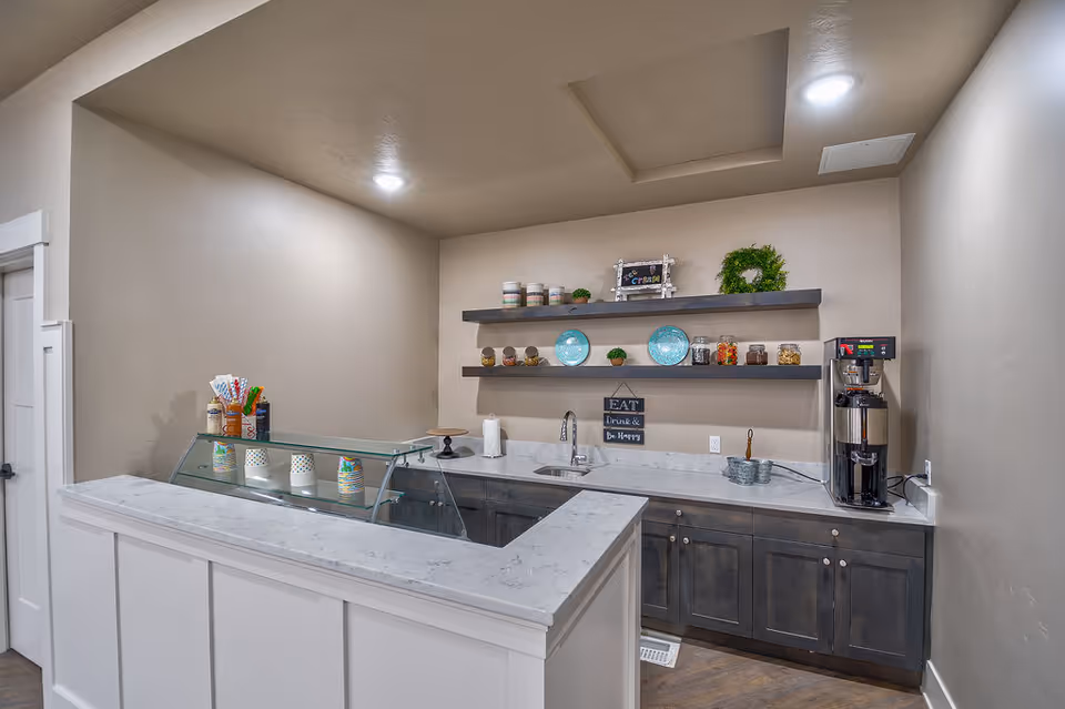 A small kitchen or snack bar area with a marble countertop, dark cabinets, a sink, and a coffee machine. Two floating shelves on the wall hold decorative plates, jars with snacks, small plants, and a wreath. There is a sign on the wall that reads 'EAT Drink & Be Happy'.