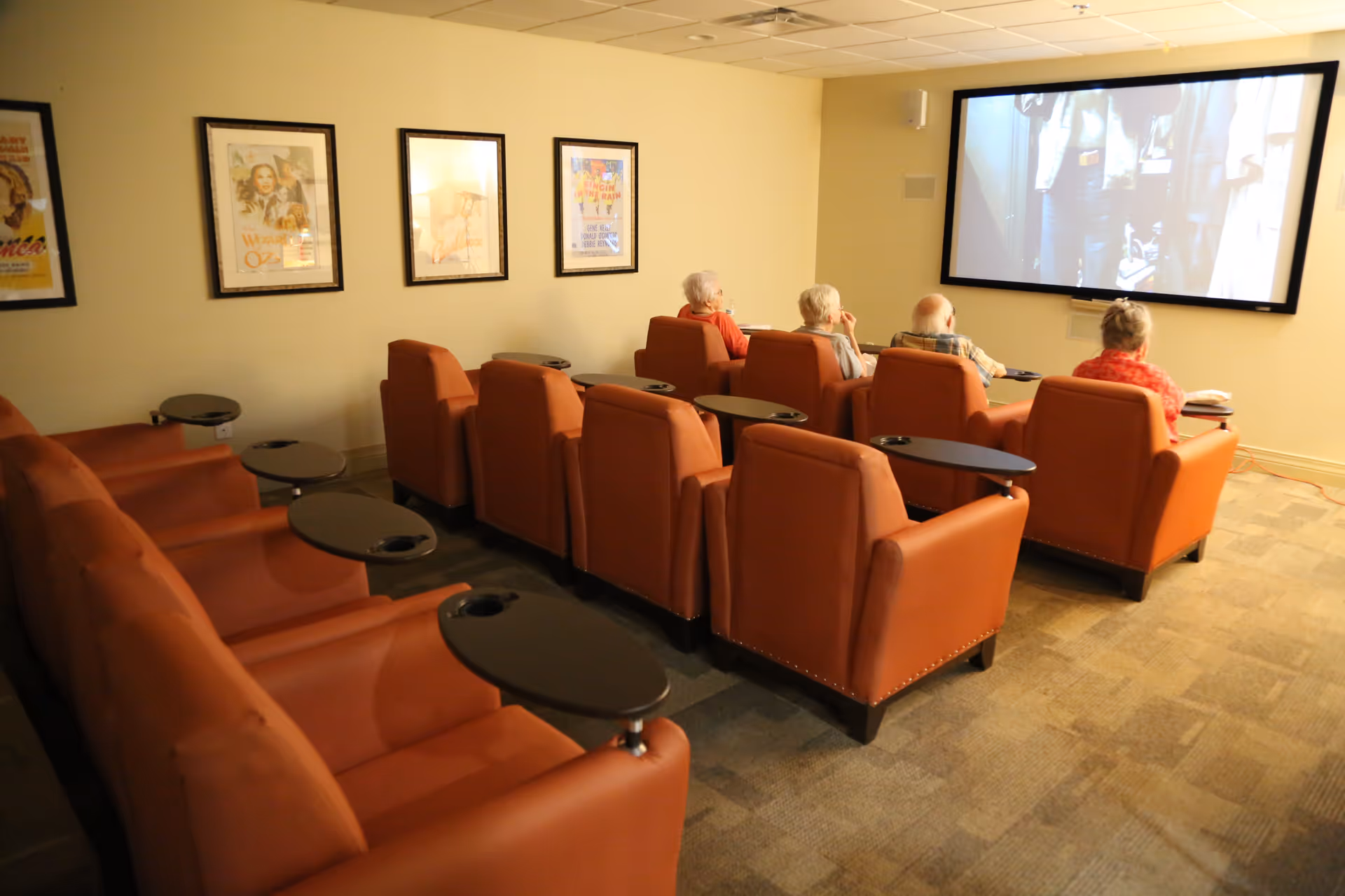 Several elderly people seated in orange recliner chairs watch a large screen in a small movie-theater style room.