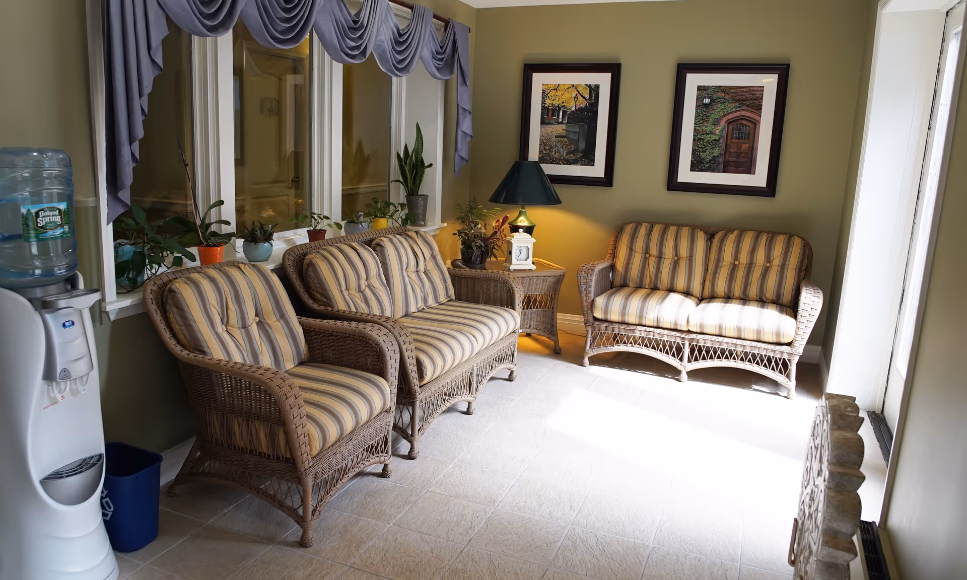 A cozy sitting area with three wicker chairs and loveseats featuring striped cushions. The room has light green walls, two framed pictures, a small wicker side table with a lamp and a clock, several potted plants on the windowsill, and a water cooler on the left side.