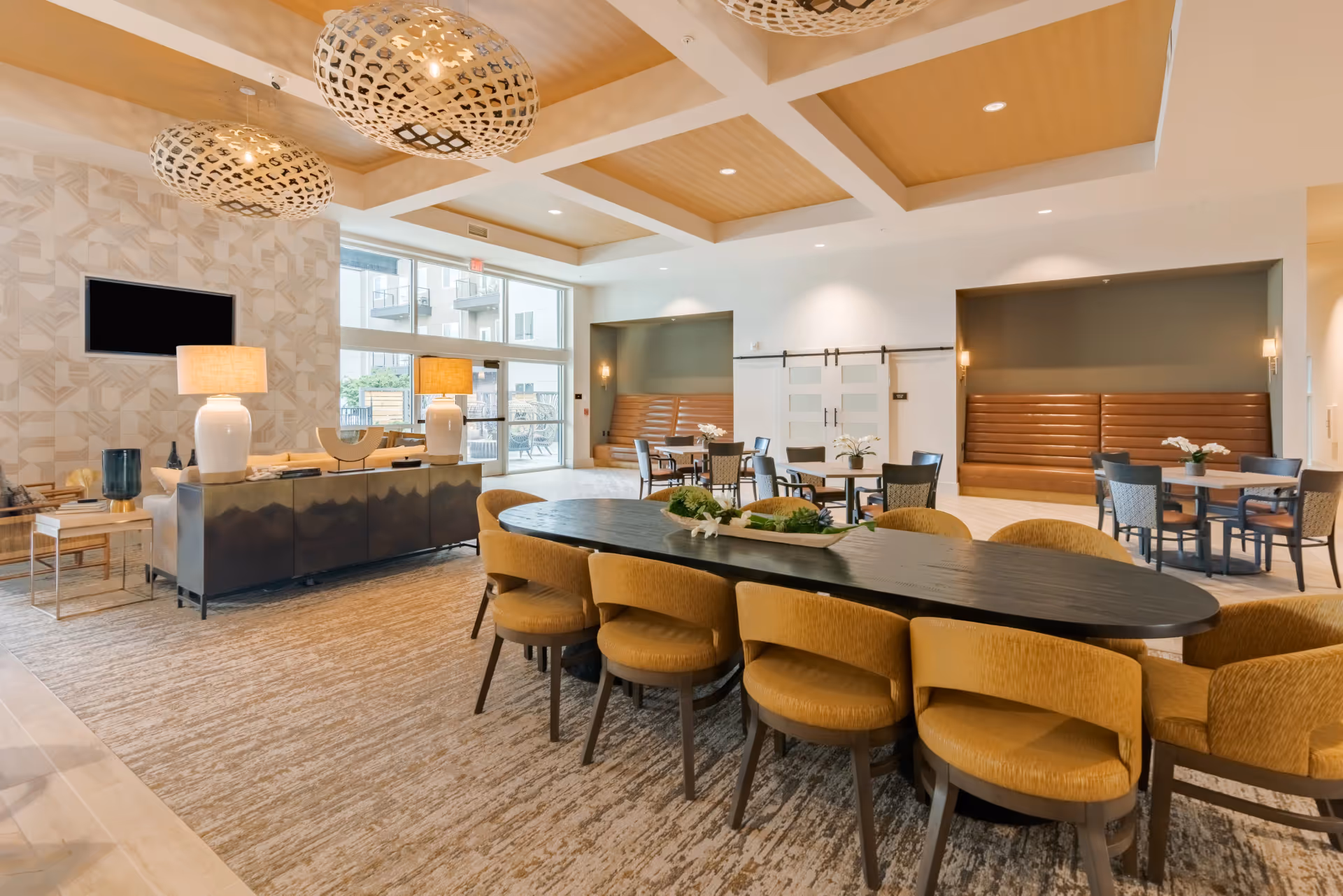 A spacious and modern common area with a large black oval table surrounded by yellow upholstered chairs in the foreground. The room features a beige patterned carpet, a seating area with a sofa and two large white lamps, and additional tables with chairs and built-in bench seating along the walls. Large windows and glass doors allow natural light to fill the space, and two decorative pendant lights hang from the ceiling.