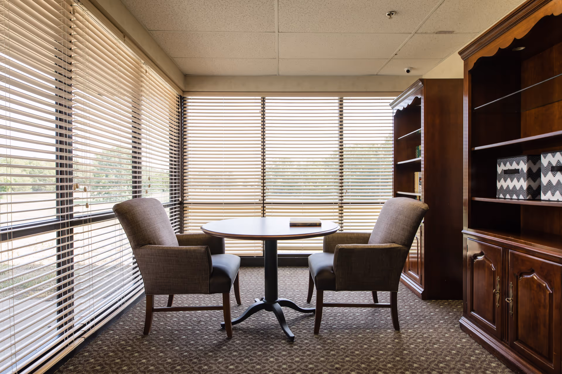 A small seating area with two upholstered armchairs facing each other across a round wooden table, positioned in front of large windows with horizontal blinds. To the right, there are wooden bookshelves with decorative storage boxes. The room has a carpeted floor and a drop ceiling with a security camera visible.