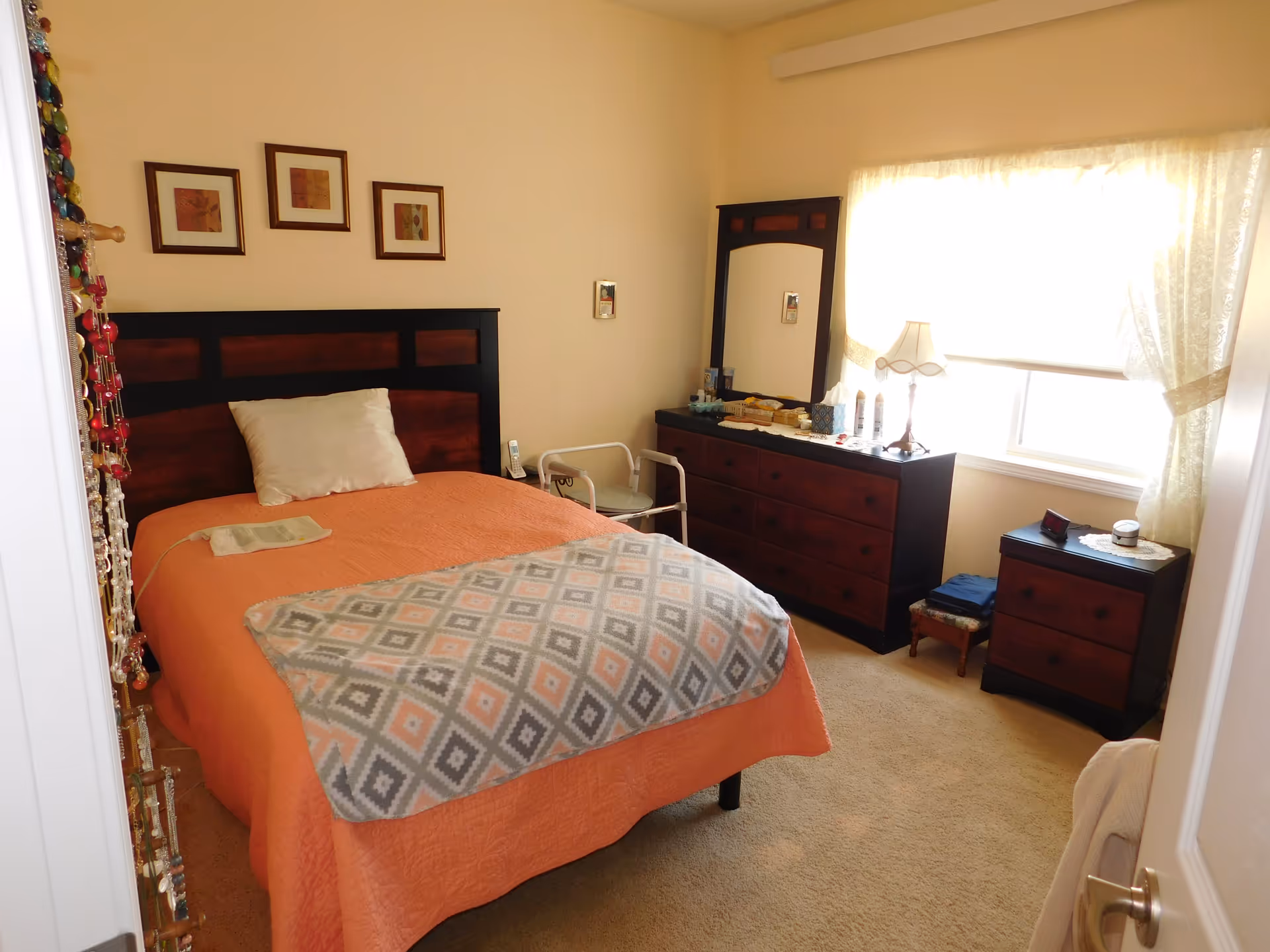 Sunlit bedroom with an orange-covered bed, dresser with mirror and lamp, nightstand, and a walker beside the bed.