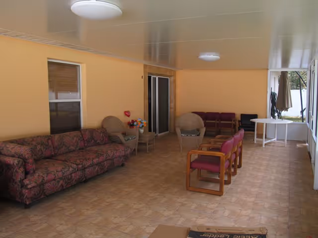 Covered indoor seating area with a patterned sofa, wicker chairs and a row of wooden-framed chairs on a tiled floor against pale yellow walls.