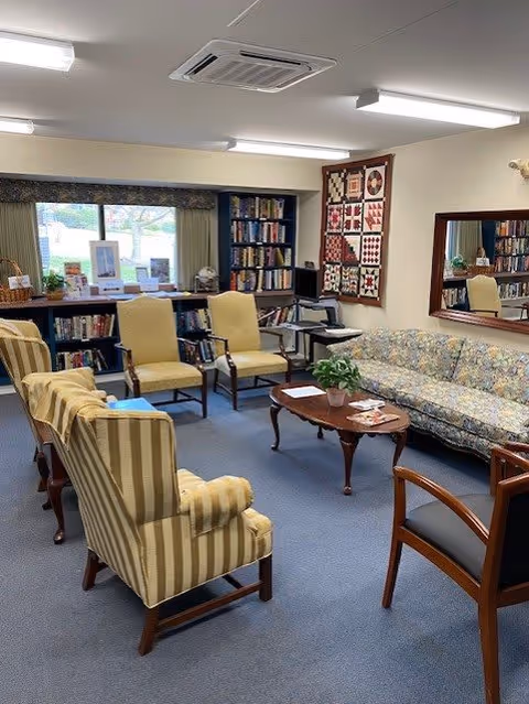 A cozy living room area with a floral patterned sofa, two striped armchairs, two yellow upholstered chairs, and a wooden coffee table with a plant and some papers on it. There is a bookshelf filled with books, a window with a valance, a quilt hanging on the wall, and a large mirror reflecting part of the room.