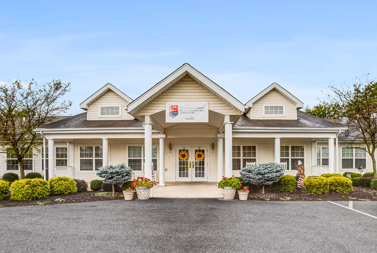 Front exterior view of a single-story senior living facility building with a covered entrance supported by white columns. The building has beige siding, multiple windows, and decorative plants and shrubs along the front. Two wreaths hang on the double glass doors under the entrance roof. A sign is displayed above the entrance.