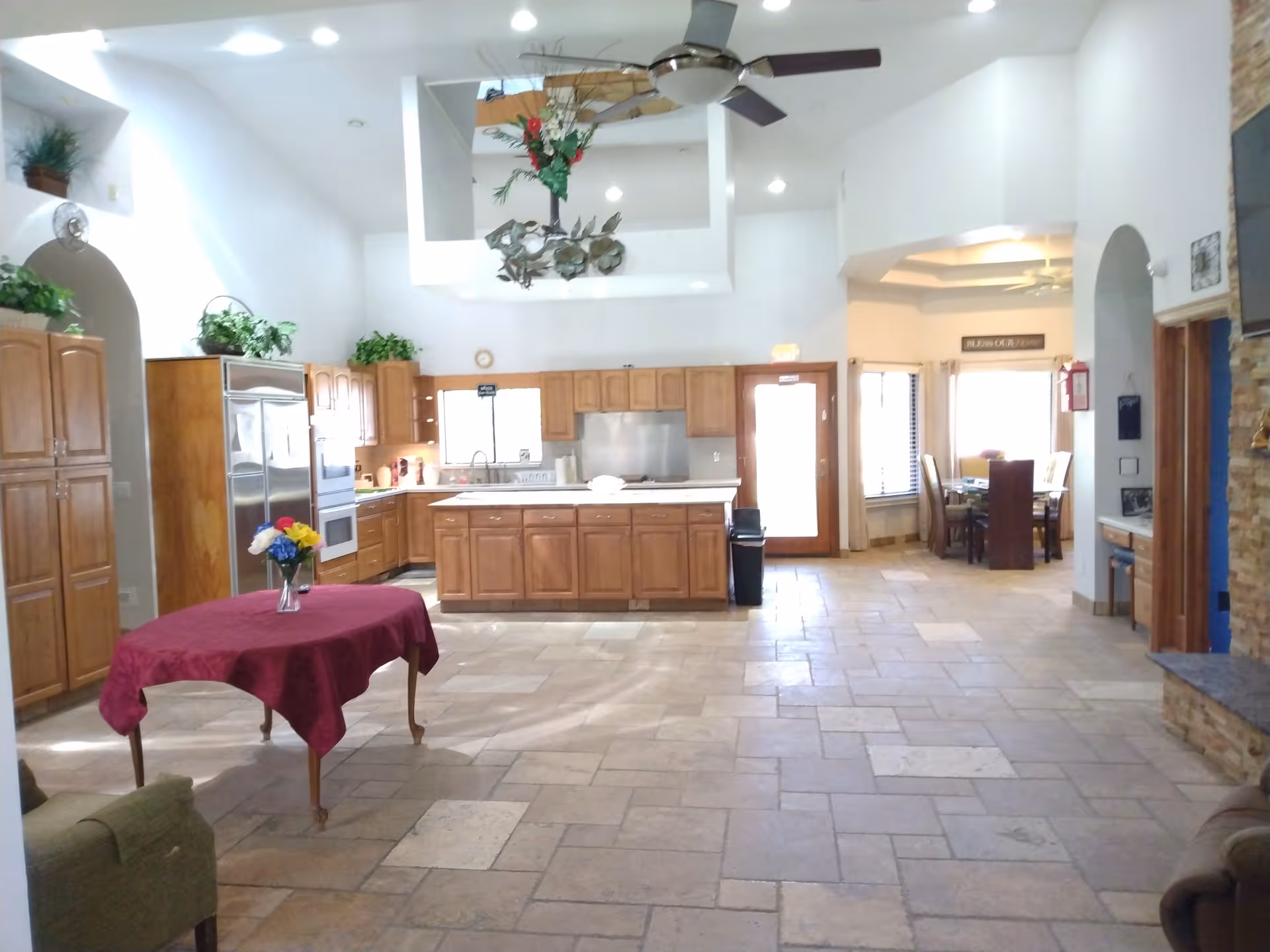 Spacious open kitchen and dining area with wooden cabinets, a central island, and a table covered with a red tablecloth holding a vase of colorful flowers. The room has tiled floors, high ceilings with a ceiling fan, and large windows letting in natural light. There is a dining table with chairs in an adjacent nook and a stone fireplace on the right side.