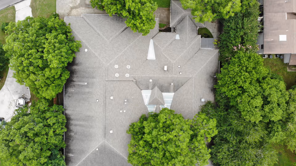 Aerial view of a building roof surrounded by green trees and adjacent structures, showing the layout and roofing details of the facility.