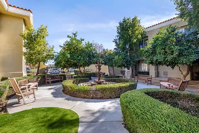 Outdoor courtyard area at Bayshire Rancho Mirage featuring a circular hedge with a central water fountain, surrounded by benches and trees under a clear blue sky.