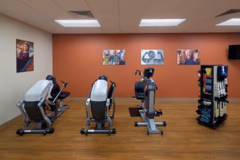 A senior fitness room with three exercise machines placed on a wooden floor facing an orange accent wall. The wall features four framed photos of elderly people engaged in various activities. To the right, there is a rack holding exercise equipment such as resistance bands and weights. The ceiling has recessed lighting panels.