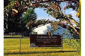 Outdoor view of a sign for Caring Acres Nursing and Rehab Center located in Toccoa, Georgia, surrounded by a fenced grassy area and trees with a clear blue sky in the background.