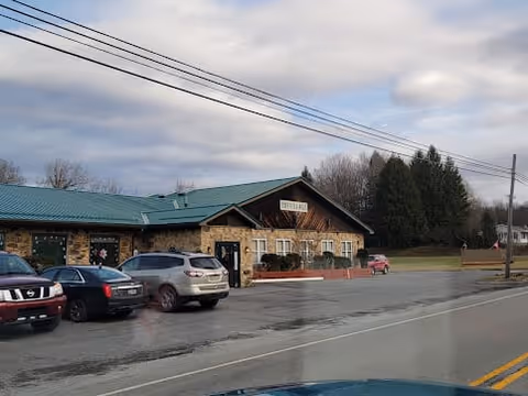 Exterior view of a single-story stone building with a green roof and a sign that reads 'The Village of Mundys Corner.' Several cars are parked in front of the building along a paved road. Trees and a cloudy sky are visible in the background.