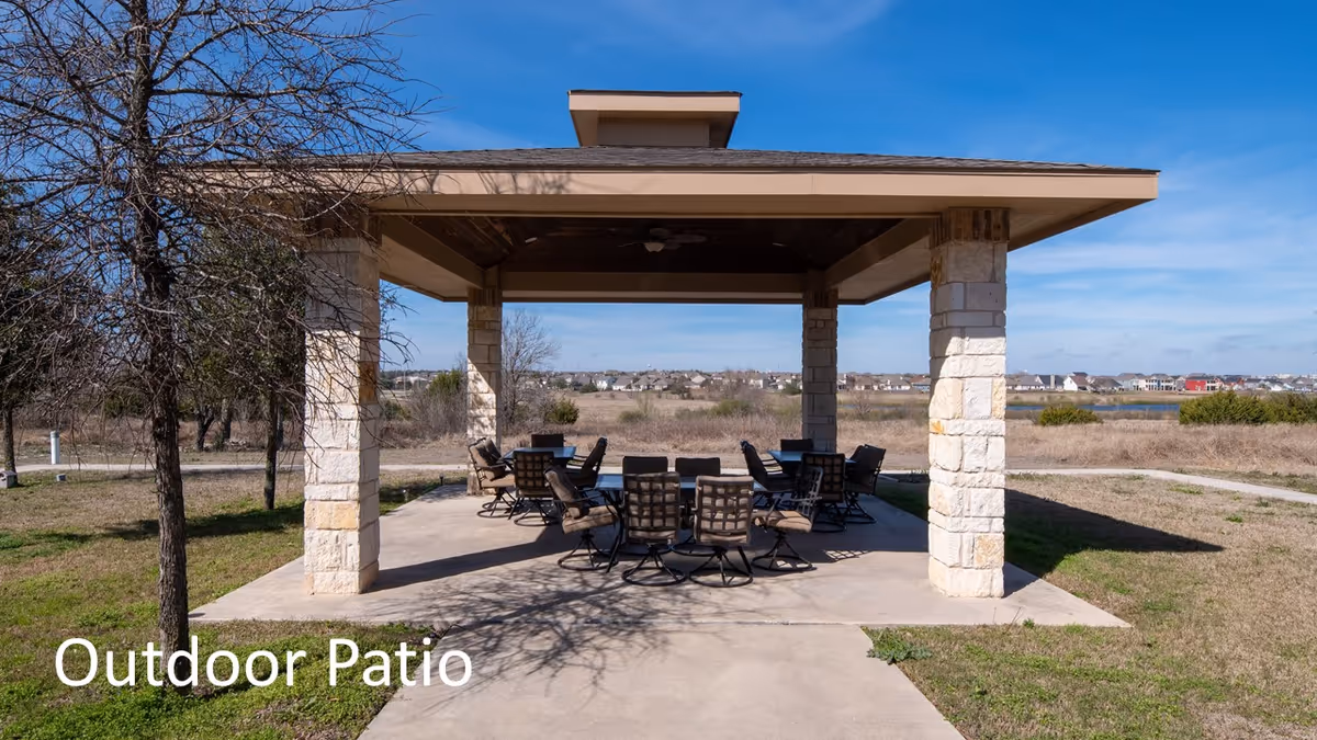 A covered outdoor patio area with stone pillars and a roof, furnished with multiple cushioned chairs and tables. The patio is set in a grassy open area with trees and a clear blue sky in the background. A walking path curves around the patio.