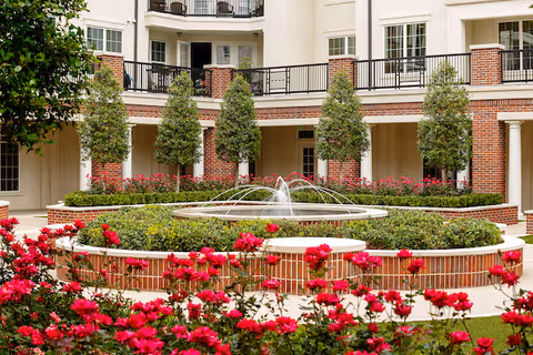 Courtyard with a circular fountain surrounded by brick planters, red flowers, and trees in front of a multi-story building.