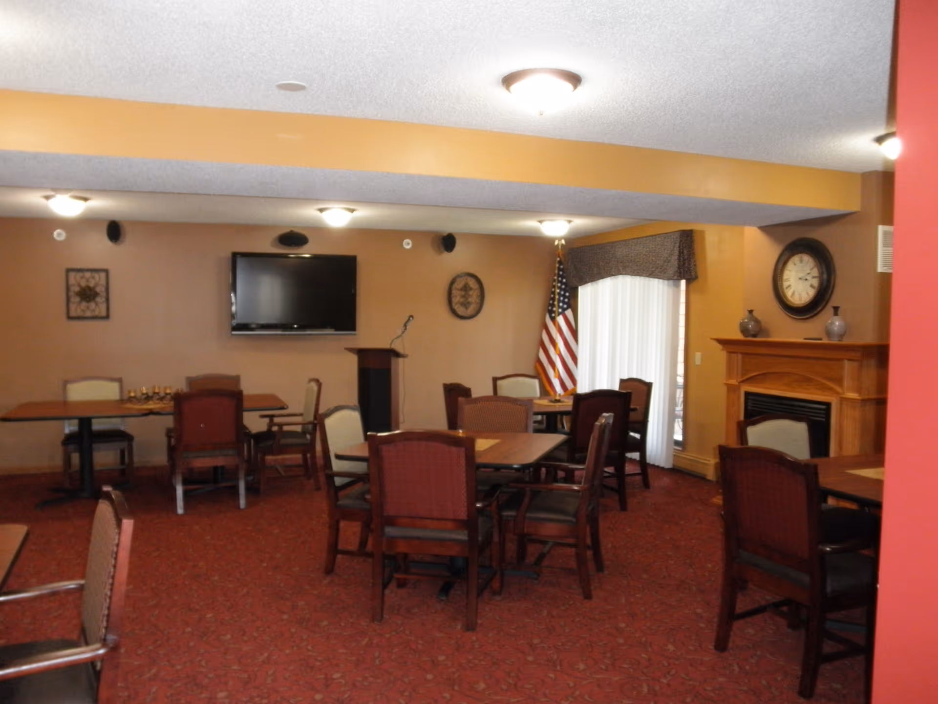 A common room with multiple tables and chairs arranged for seating. The room features a wall-mounted flat screen TV, a podium with a microphone, an American flag near a sliding glass door with vertical blinds, a fireplace with a clock and decorative vases above it, and wall decorations. The carpet is red with a subtle pattern, and the walls are painted in warm tones.