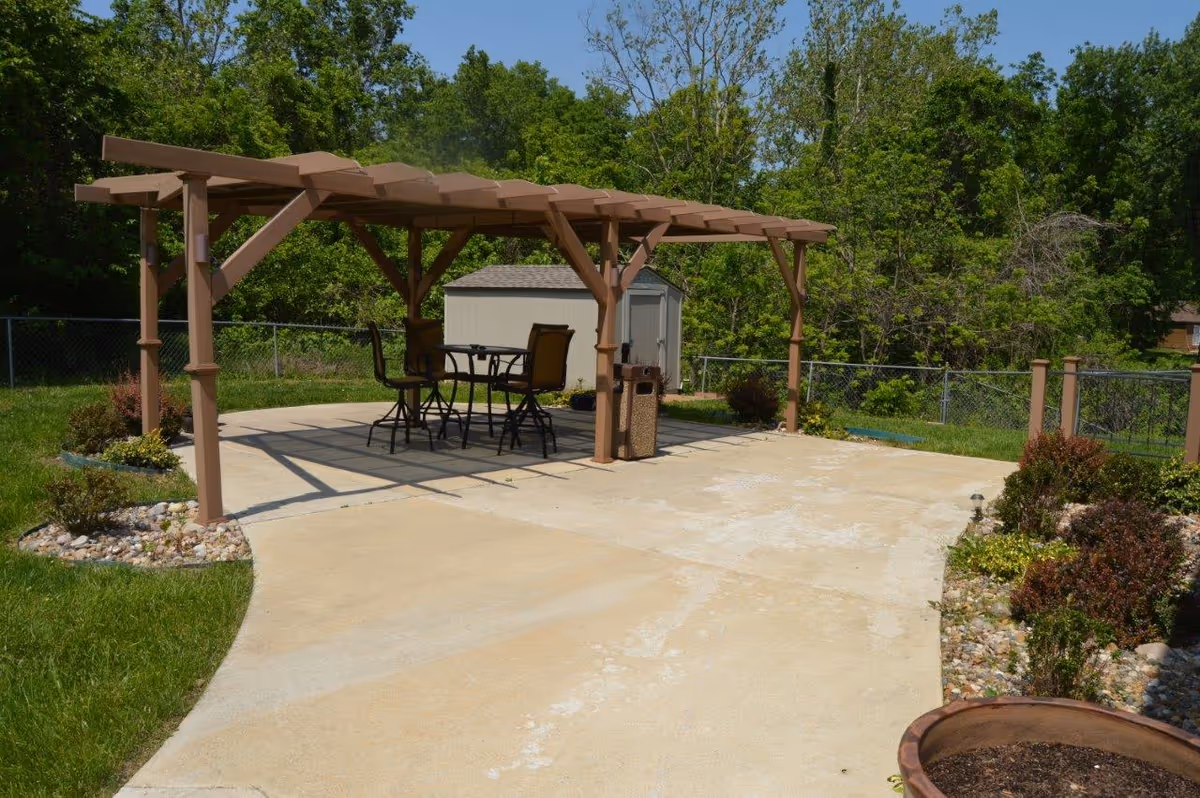 A backyard patio with a wooden pergola over a table and chairs, a small shed, and surrounding trees and landscaping.