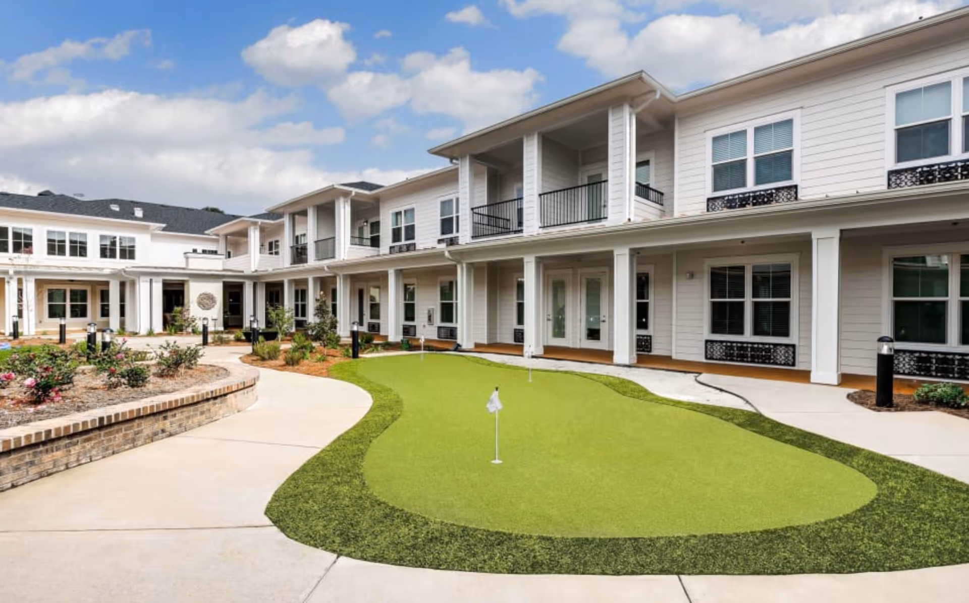 Outdoor courtyard area of The Blake at Pensacola featuring a small artificial putting green with flags, surrounded by a paved walkway and landscaped garden beds. The two-story building with white siding and multiple windows and balconies encloses the courtyard under a partly cloudy sky.