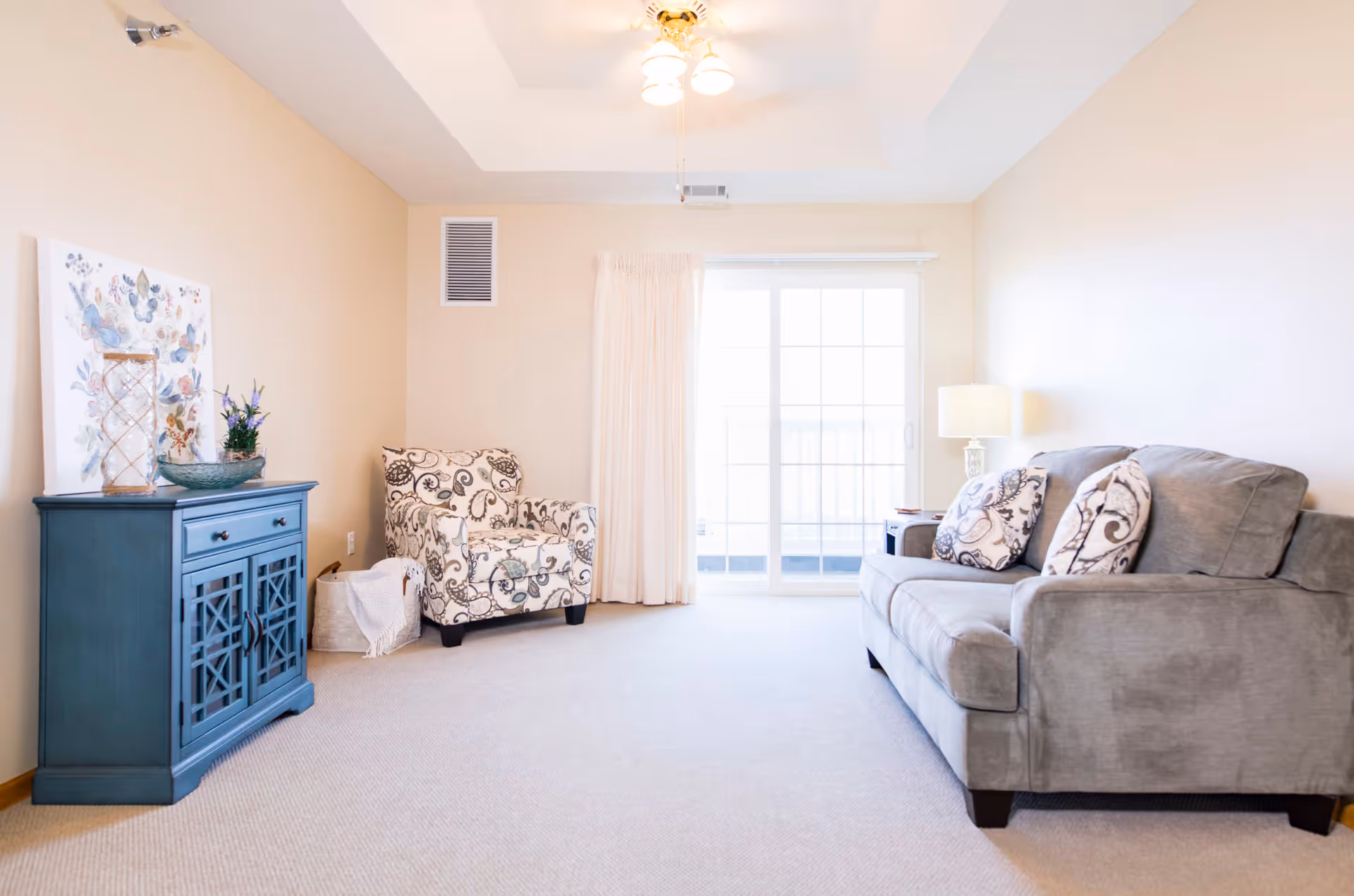 Bright living room with a gray sofa, patterned armchair, blue cabinet, and sliding glass door.