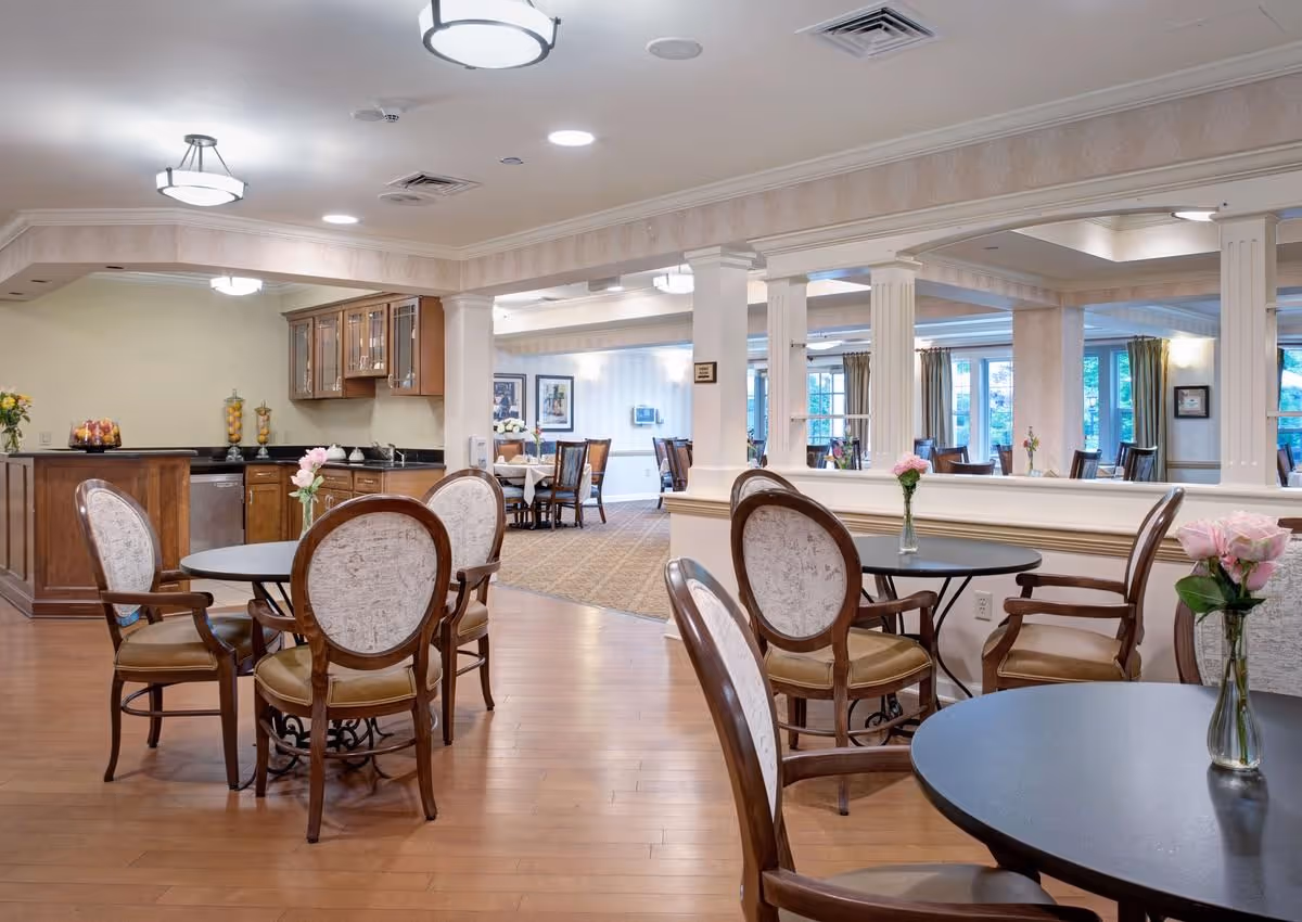 Bright communal dining area with round tables, upholstered chairs, a serving counter, and vases of flowers.
