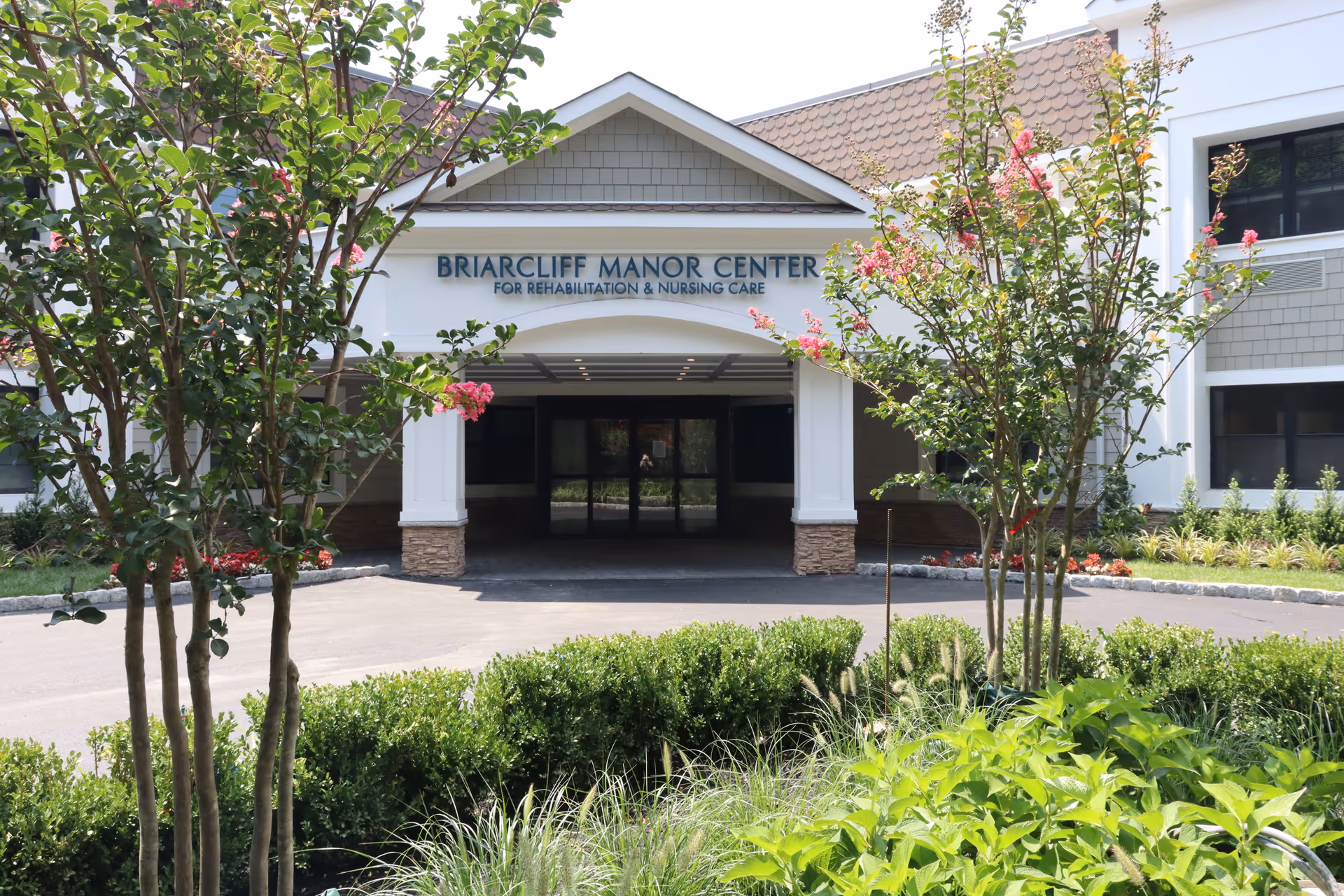Front exterior view of Briarcliff Manor Center for Rehabilitation and Nursing Care building with a covered entrance, surrounded by landscaped greenery and flowering trees.