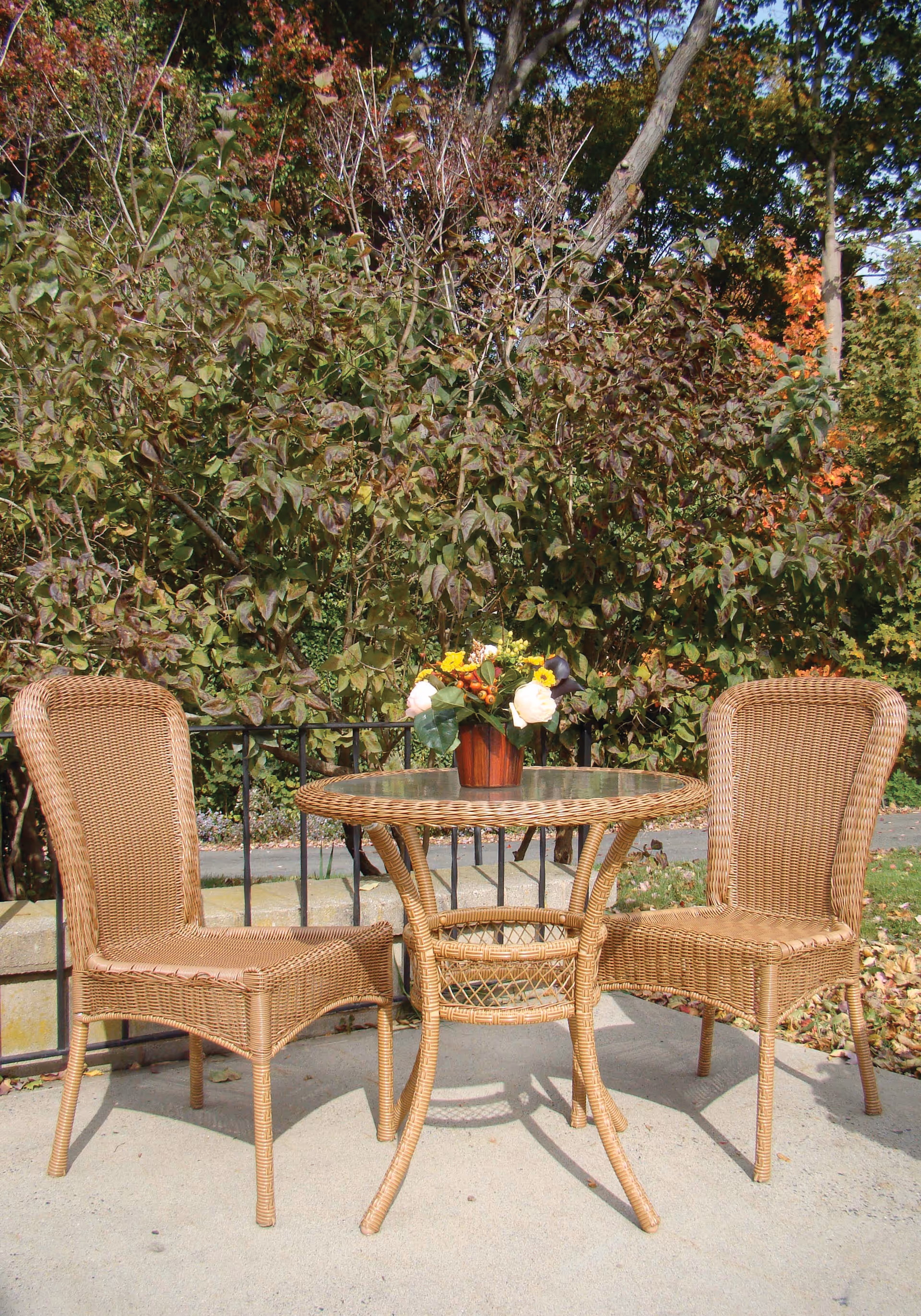 Outdoor patio area with a round wicker table and two matching wicker chairs. A small flower arrangement in a brown pot is placed on the table. Behind the patio is a black metal railing and dense green foliage with some autumn-colored leaves.